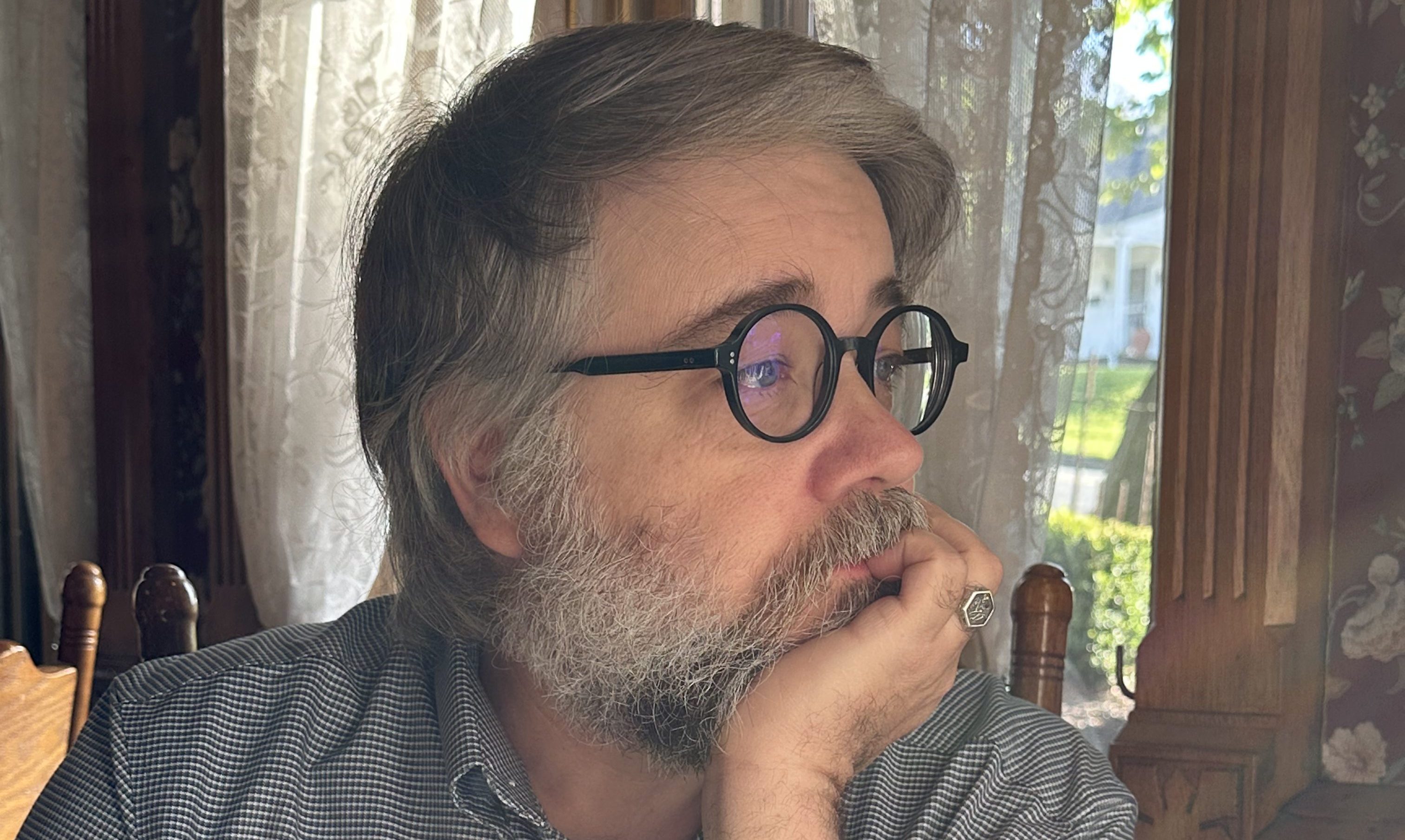 Author Derek Maxfield looking to the left with head in hand. Maxfield has grey hair parted to the side, a short trimmed grey beard, and round black-rimmed glasses. 